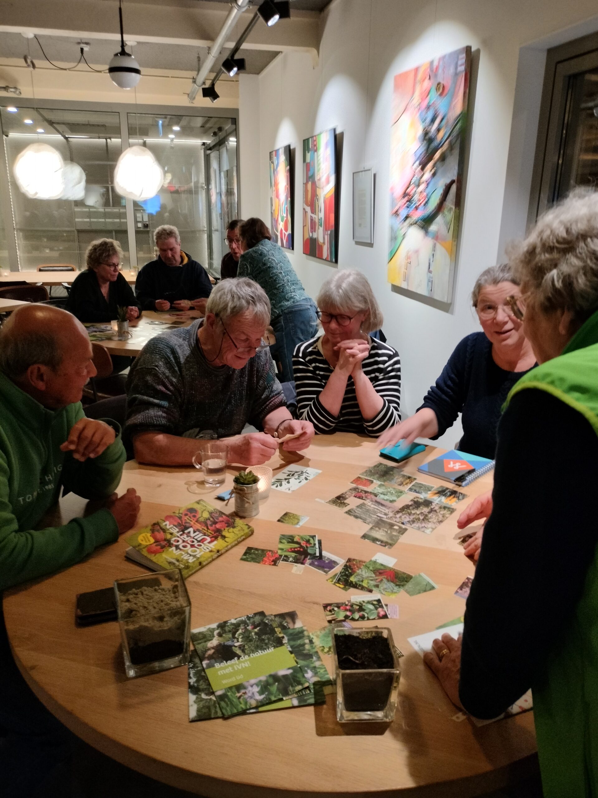 Mensen aan tafel bespreken tuinbouw, met plantenfoto's en boeken op tafel in een moderne ruimte.