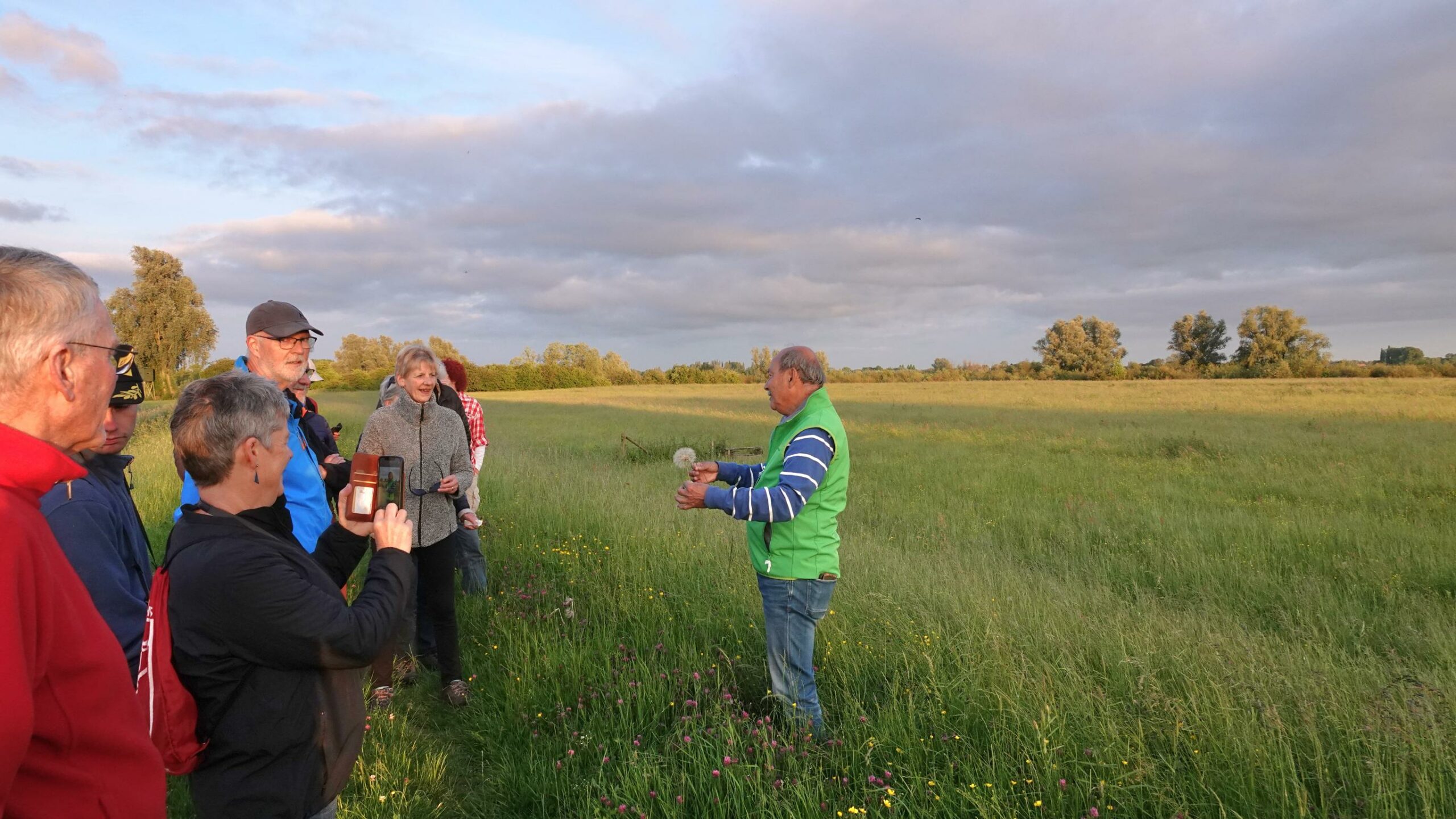 Een groep mensen volgt een buitenpresentatie in een groen veld met een bewolkte lucht.
