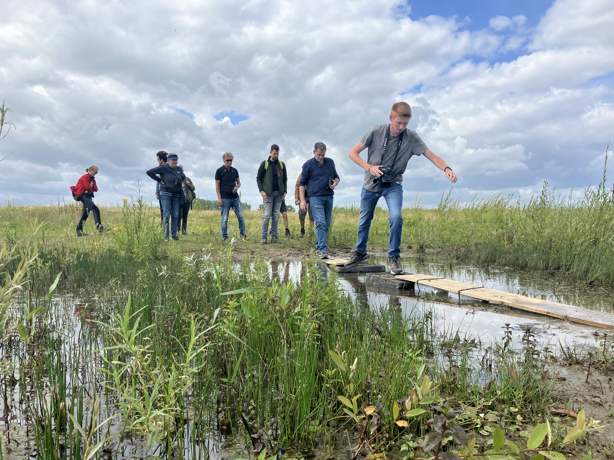 Groep mensen steekt smalle houten brug over in grasrijke omgeving onder bewolkte lucht.