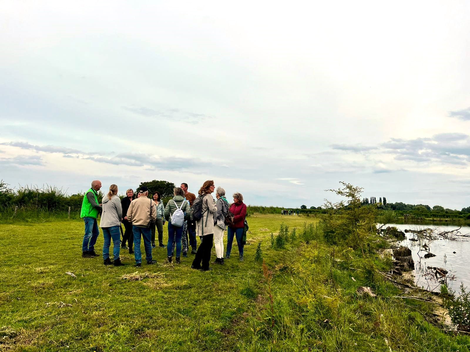 Groep mensen staat in een open veld bij een rivier, omgeven door natuur en bewolkte lucht.