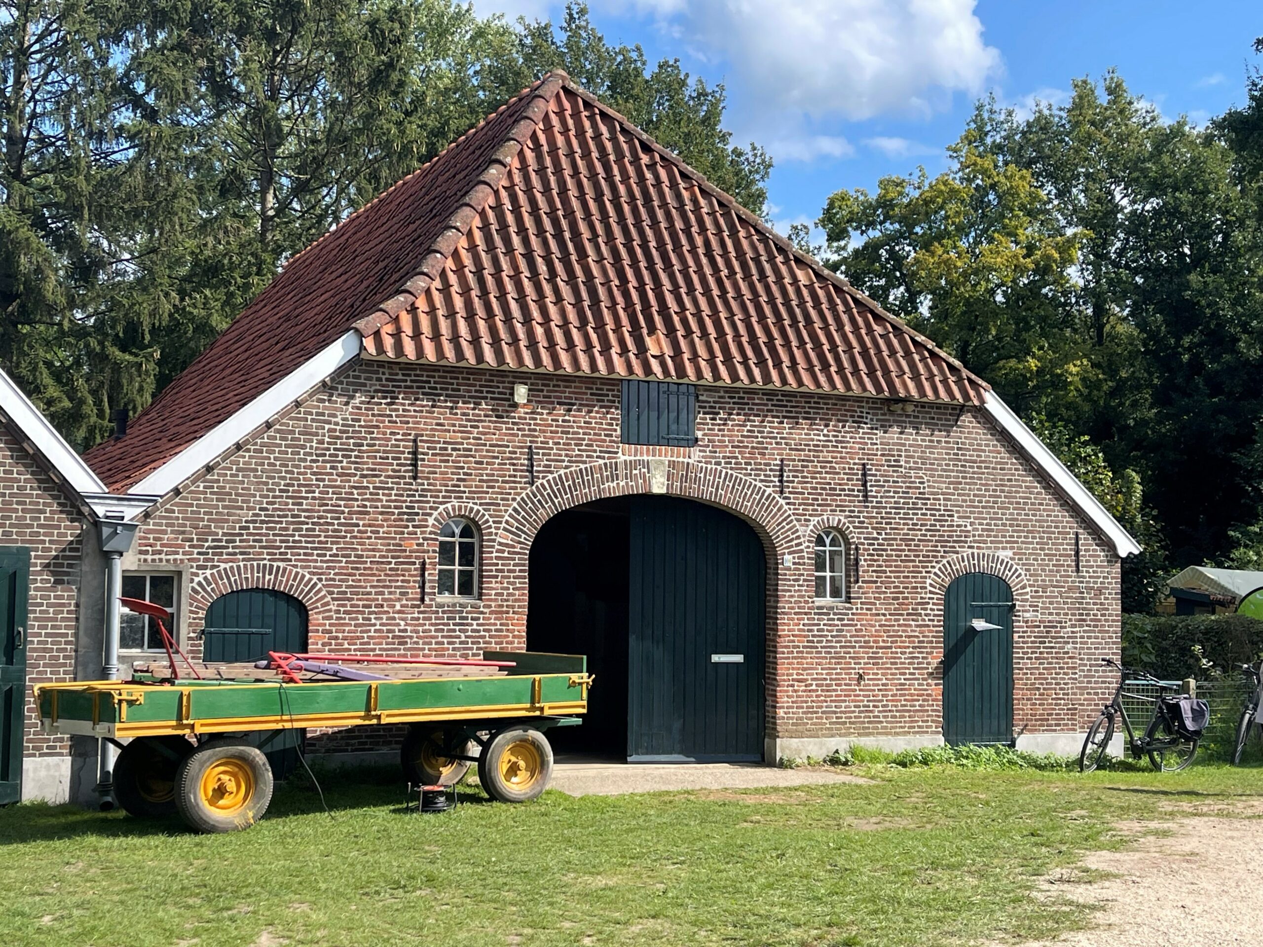 Oude bakstenen schuur met rood dak, groene poort, wagen en fiets ervoor, omringd door bomen.