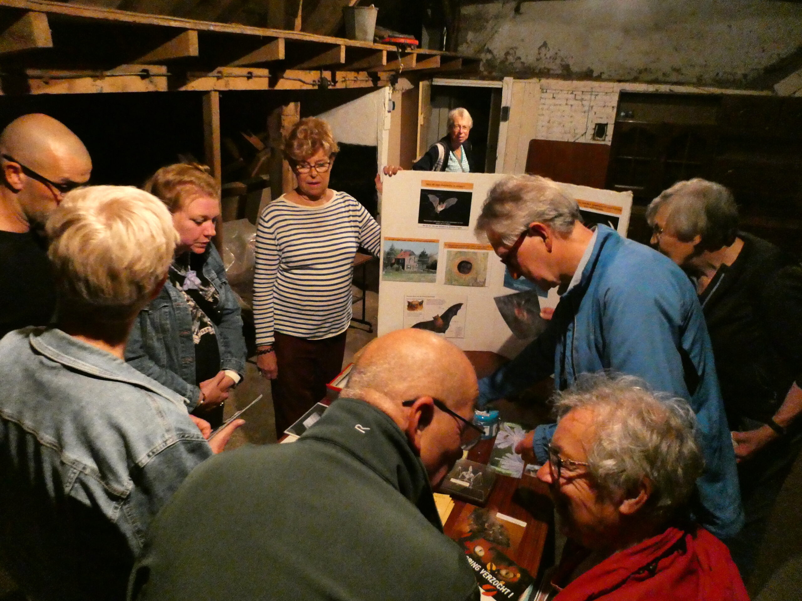 Groep mensen bekijkt natuurfoto's en boeken in een donkere ruimte. Een persoon wijst op een bord.
