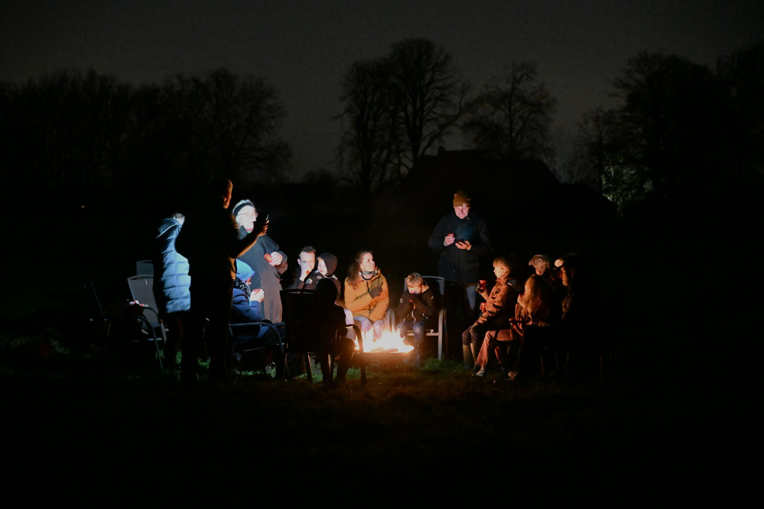 Mensen zitten 's nachts in een kring rond een kampvuur, omringd door donkere bomen.