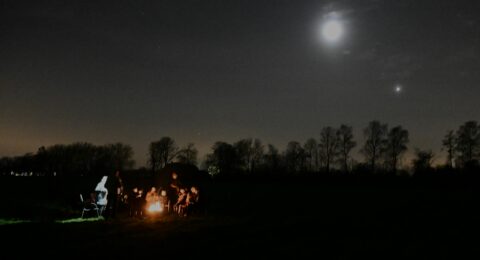 Een groep mensen zit 's nachts rond een kampvuur onder een heldere maan.