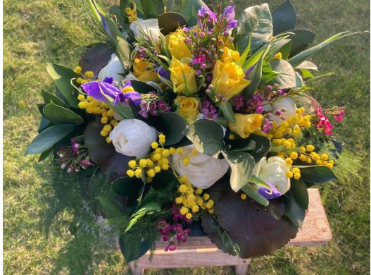 Boeket met gele rozen, witte pioenrozen, paarse irissen en groen blad op een houten tafel.