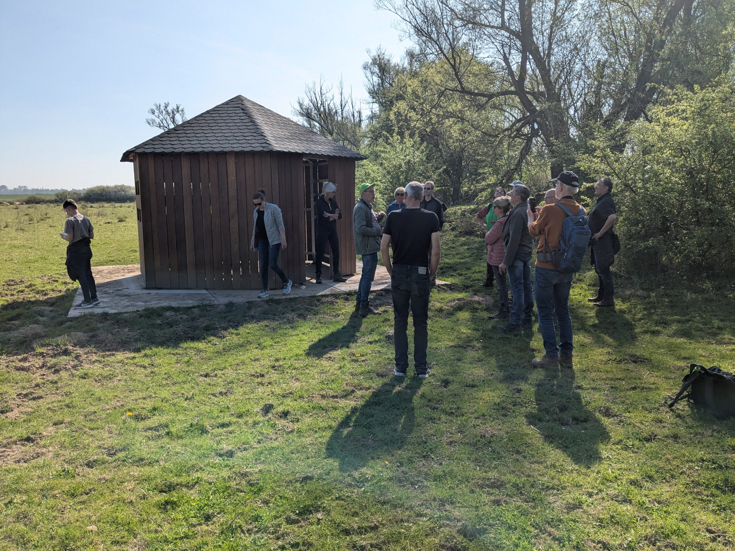 Een groep mensen staat buiten bij een houten gebouw in een groene, bosrijke omgeving.