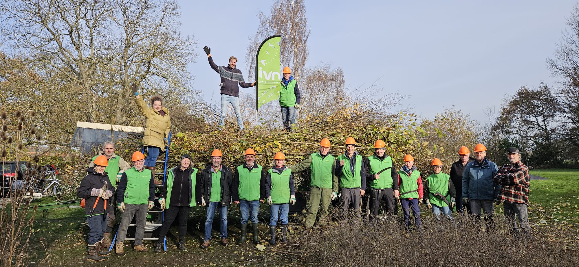 Groep vrijwilligers in groene hesjes en oranje helmen bij een stapel takken, met een IVN-vlag.