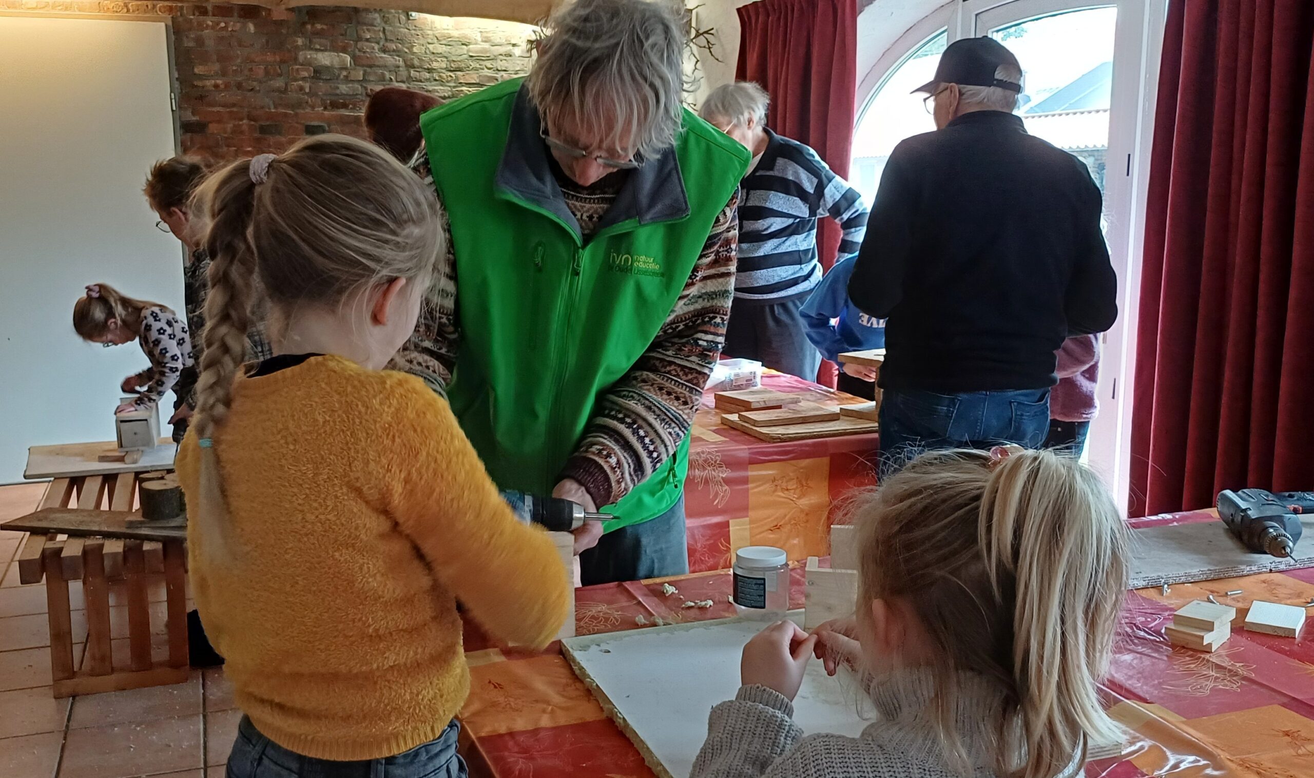 Kinderen en volwassenen knutselen samen aan een tafel met hout en gereedschap.