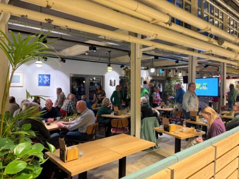 Groepsbijeenkomst in een café-achtige ruimte met mensen aan tafels en kunst aan de muur.