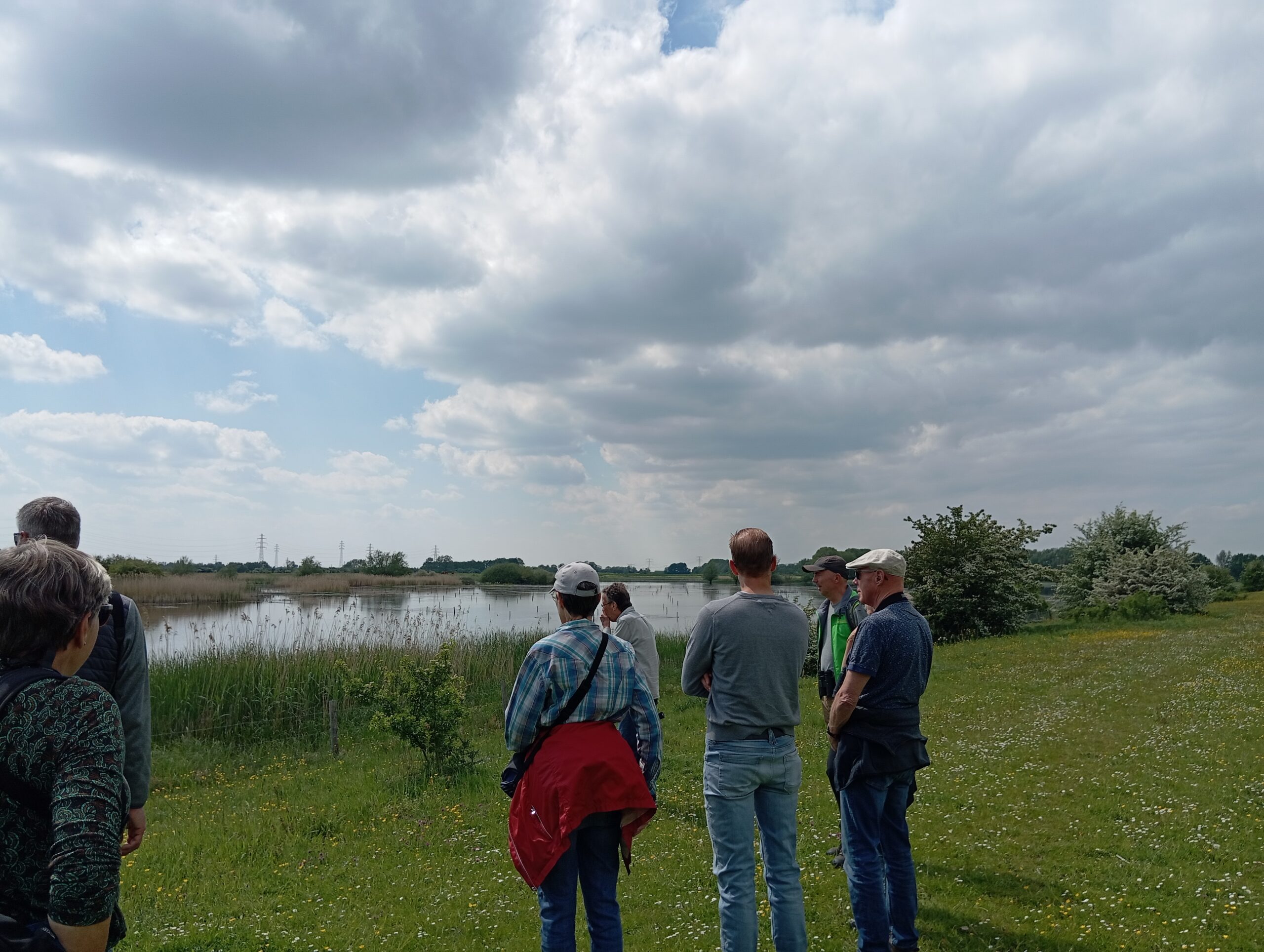 Een groep mensen kijkt uit over een meer met wolkenluchten boven een groene oever.