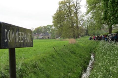 Een bord met "DOMINEESPAD" bij een grasveld, met een groep mensen en een boerderij op de achtergrond.