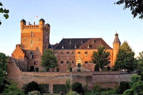 Middeleeuws kasteel met torens, omgeven door groene bomen en een stenen muur bij zonsondergang.