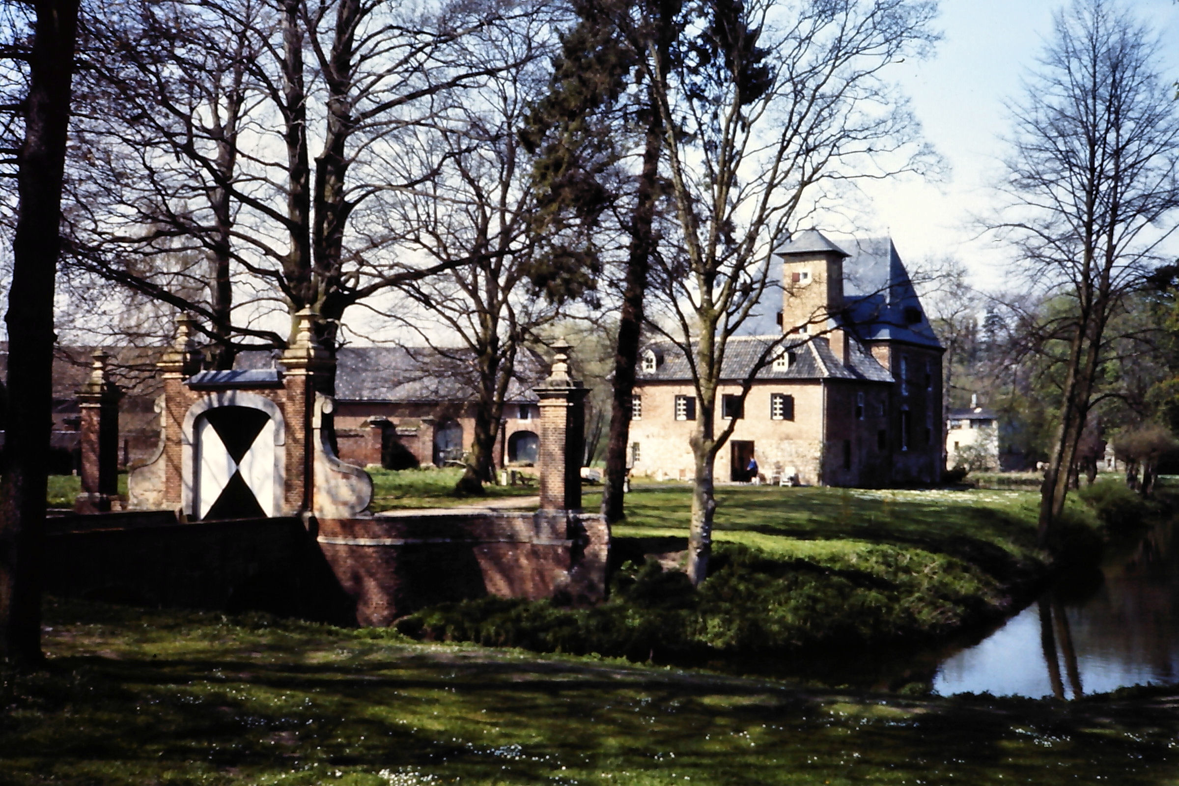 Historisch kasteel met toren en gracht, omgeven door bomen en een poort in een groene omgeving.