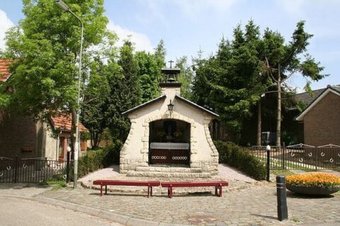 Kleine stenen kapel met kruis, omgeven door bomen, met bankje aan de voorkant.