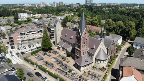 Luchtfoto van een kerk met kerktoren omringd door begraafplaats, woningen en bomen.