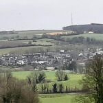 Landelijk landschap met een dorp, omgeven door glooiende groene heuvels en verspreide bomen.