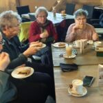 Een groep mensen geniet van koffie en gebak rond een houten tafel in een café.