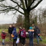 Een groep mensen luistert naar een gids onder een grote, kale boom in een bosrijke omgeving.