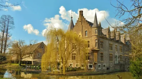 Kasteel aan een gracht met een wilgenboom en terras, op een zonnige dag met blauwe lucht en wolken.