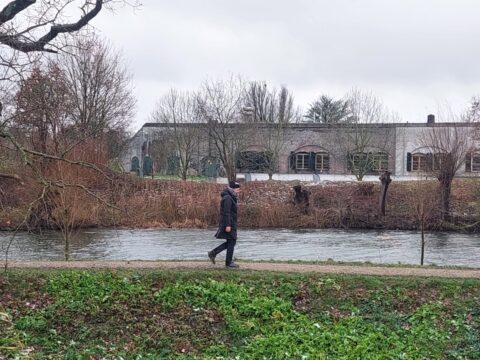 Persoon wandelt langs een rivier, met een gebouw en kale bomen op de achtergrond.
