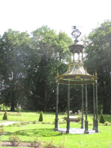 IJzeren prieel met sierlijke details in een groene tuin, omgeven door hoge bomen.