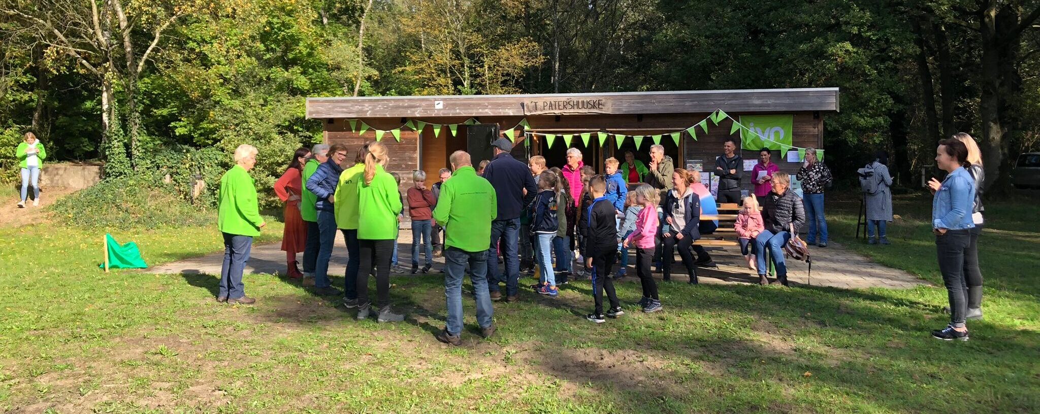 Een groep mensen verzameld bij een houten gebouw in een park met groene omgeving en zonlicht.