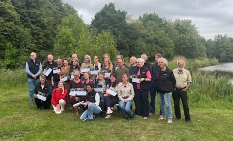 Groepsfoto van mensen in een park, lachend en poserend met certificaten, tegen een bosrijke achtergrond.