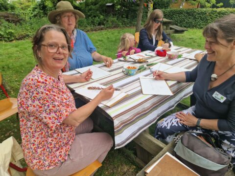 Mensen tekenen aan een tafel in een tuin, omringd door bomen.