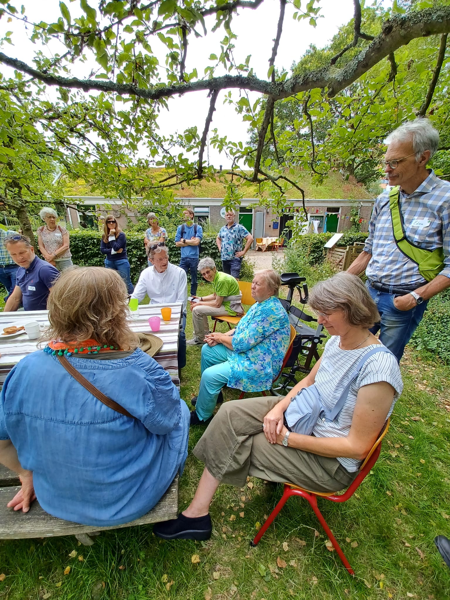 Groep mensen zit en staat rondom een tafel in een groene tuin, betrokken in een gesprek.