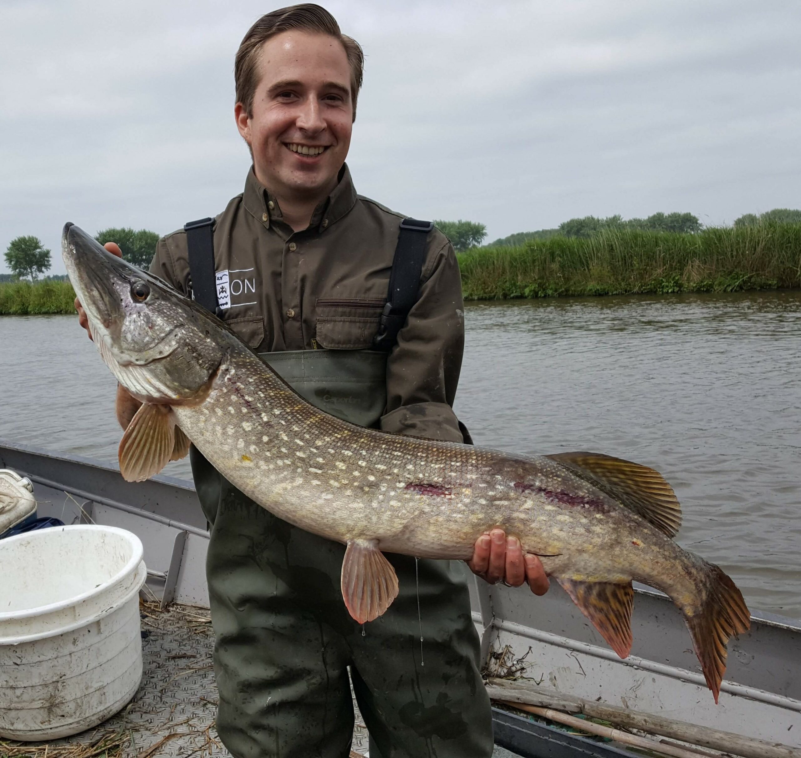 Visser poseert met grote snoek in zijn handen, staand op een boot bij een rivier.