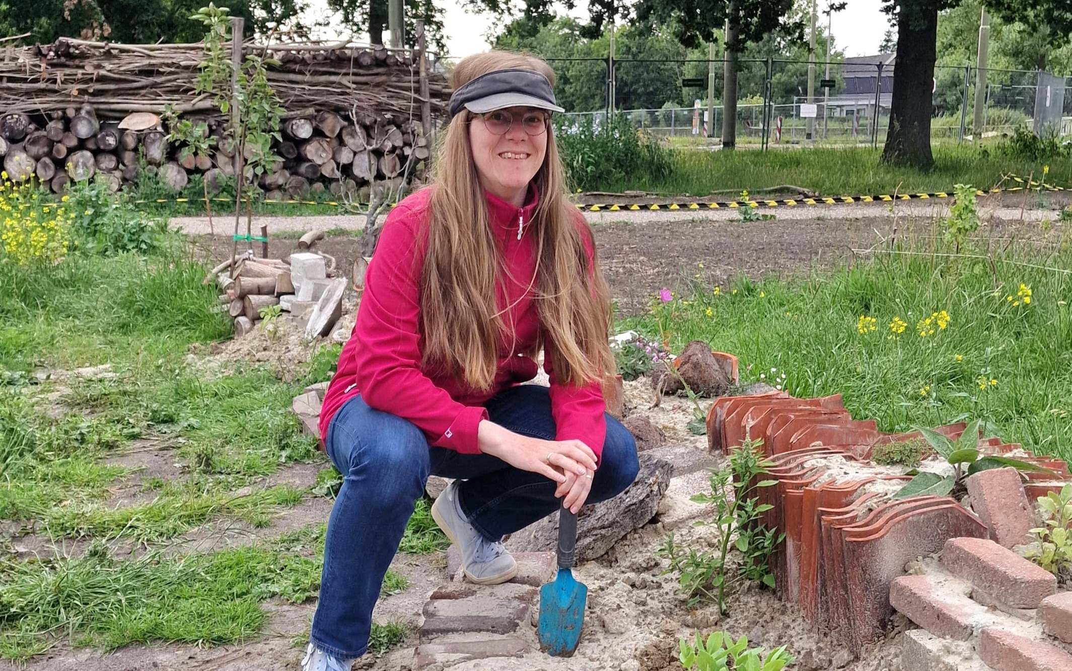 Persoon met rode jas en pet tuiniert, omgeven door groen en houtblokken.