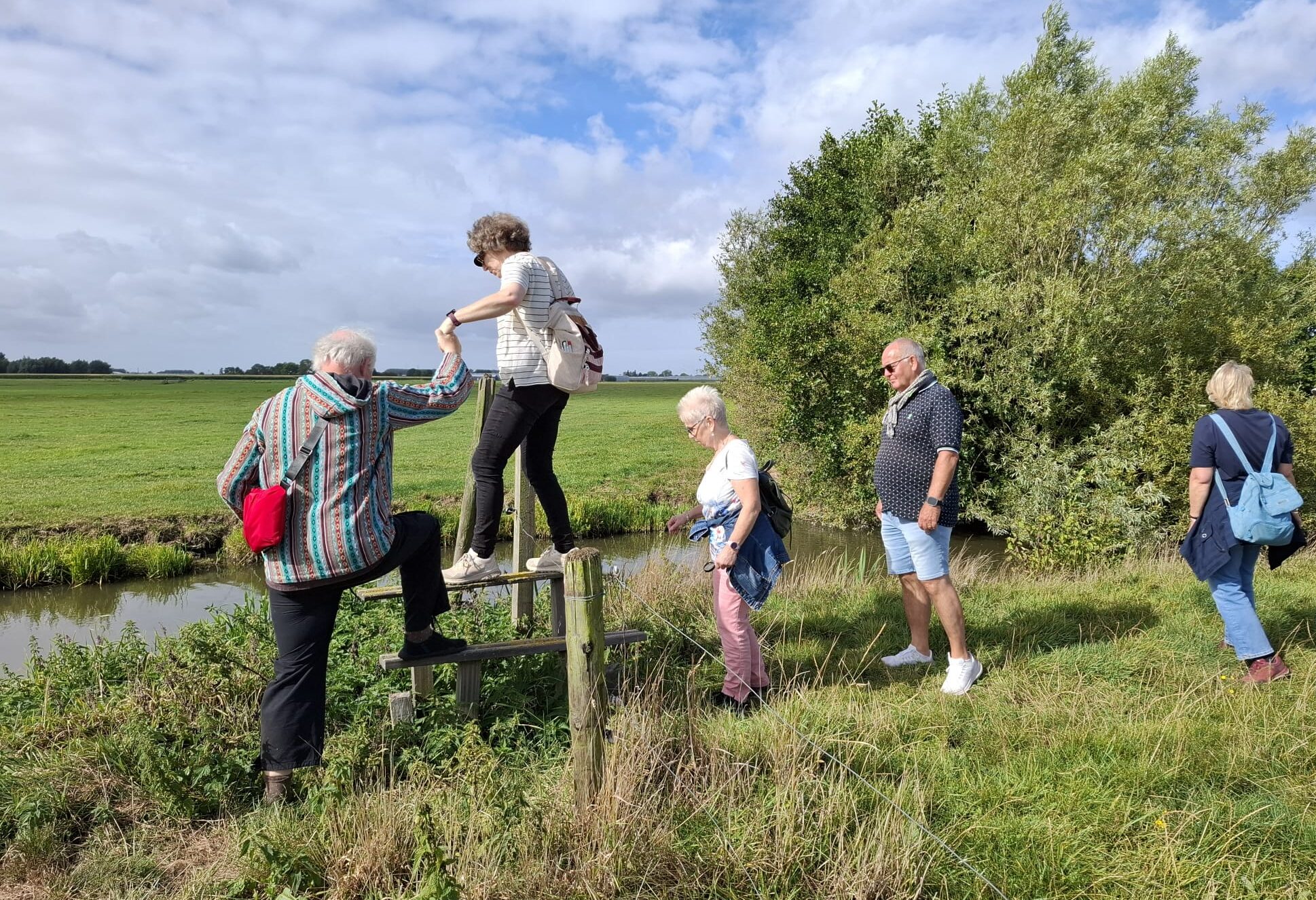 Groep wandelaars steekt een houten hek over in een groen, open landschap met water en bomen.