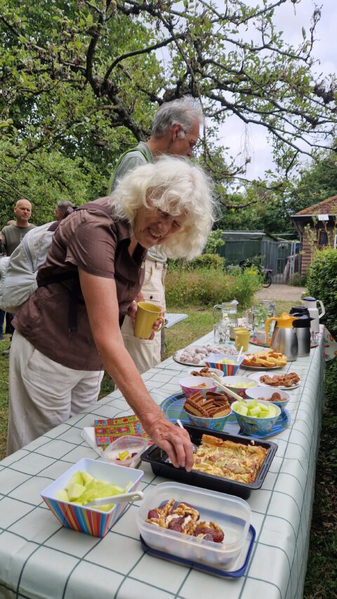 Mensen pakken eten van een tafel met snacks en drankjes in een tuinsetting.