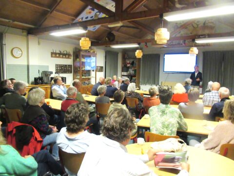Een groep mensen luistert naar een lezing in een klaslokaal met een presentator voor een scherm.