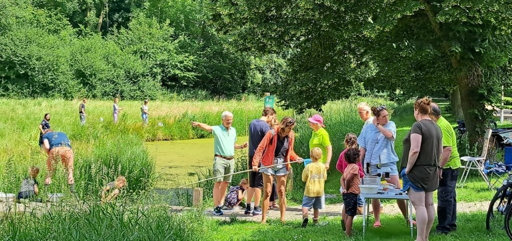 Groep mensen bij een vijver in een groen park, kinderen spelen en volwassenen praten onder een boom.