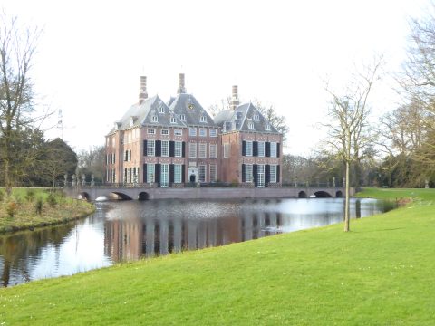 Historisch kasteel met gracht, omgeven door grasvelden en bomen onder een heldere hemel.