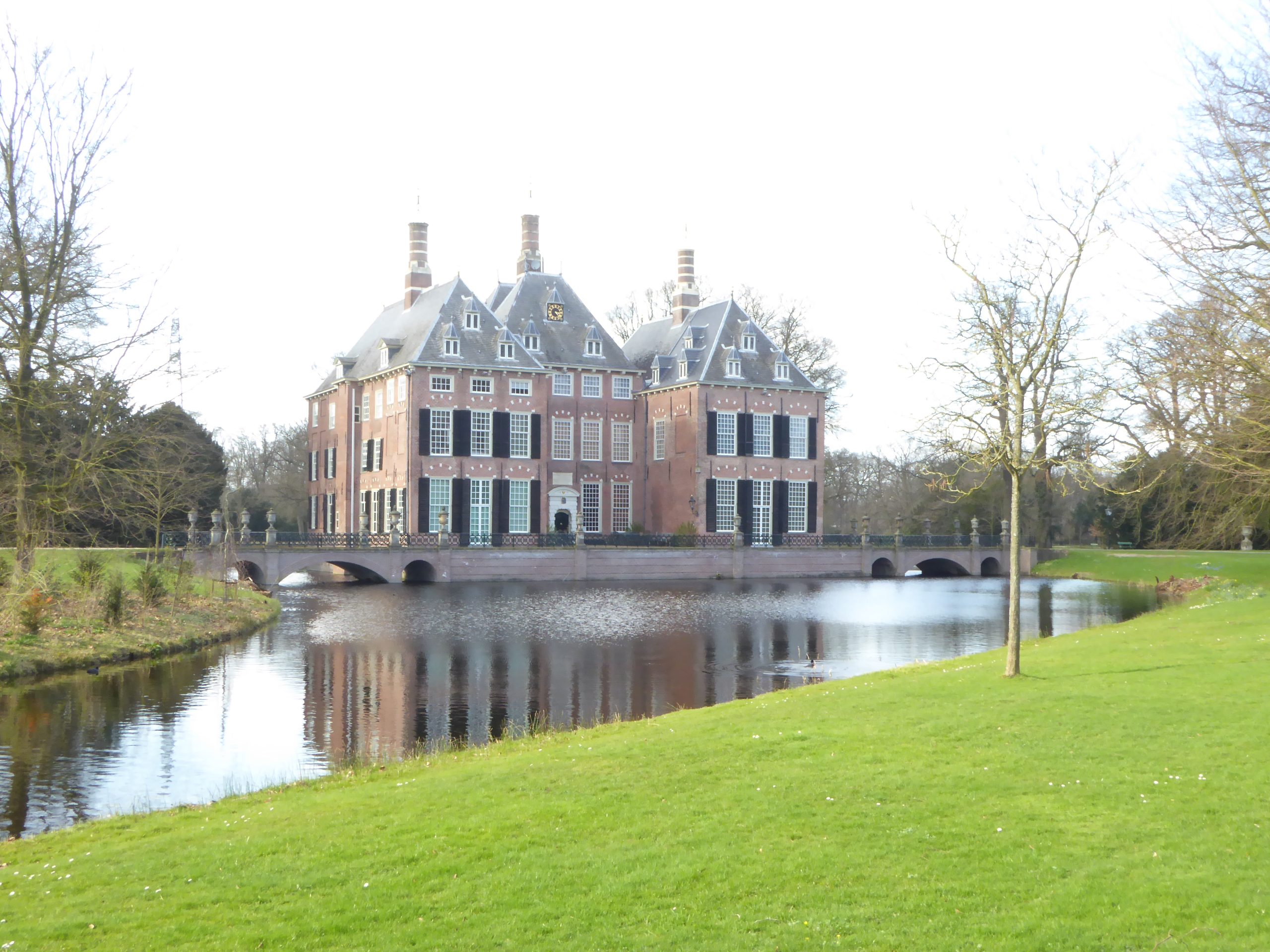 Historisch kasteel met gracht, omgeven door grasvelden en bomen onder een heldere hemel.