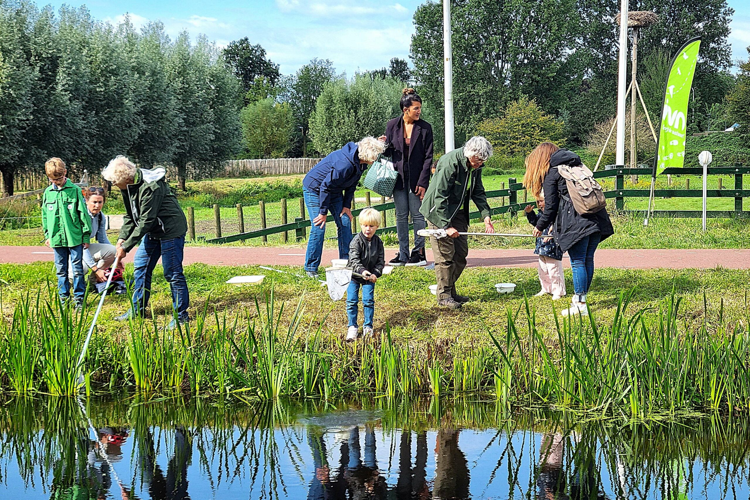 Mensen bezig met natuuractiviteiten bij een vijver, met schepnetten en groene omgeving.