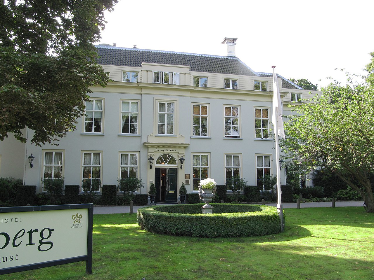 Groot herenhuis met tuin en vlag, bord met "Hotel" op de voorgrond.
