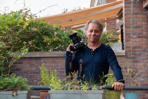 Persoon met camera staat buiten tussen planten op een balkon.