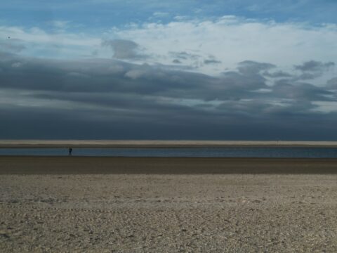 Breed strand met eenzame wandelaar en rustige zee onder dreigende, bewolkte lucht.