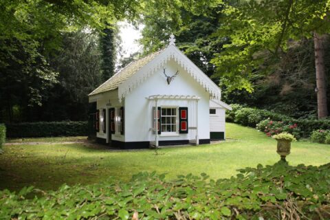 Wit huisje met hertenhoofd op gevel, omgeven door groen gazon en bosrijke omgeving.