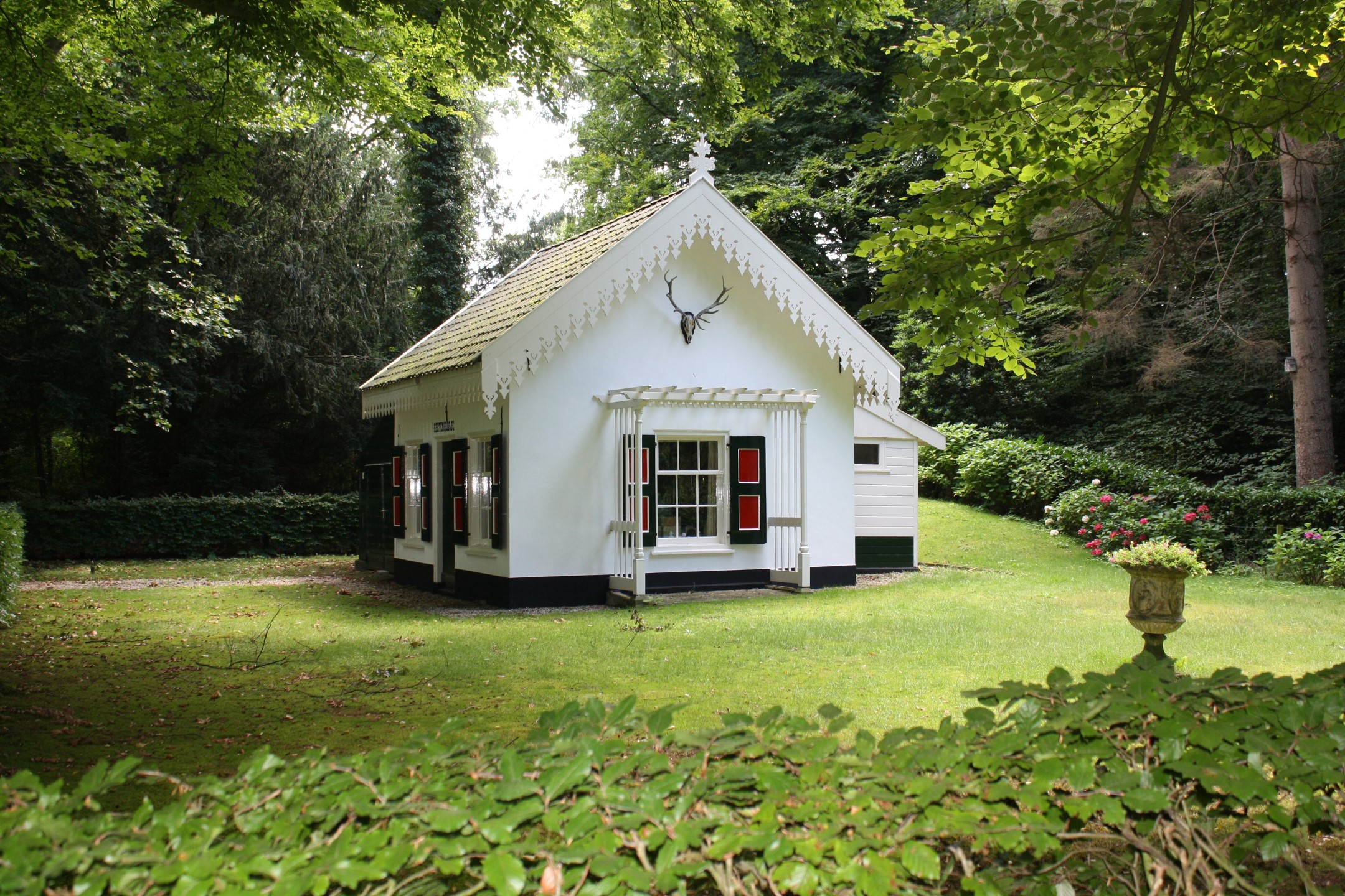 Wit huisje met hertenhoofd op gevel, omgeven door groen gazon en bosrijke omgeving.