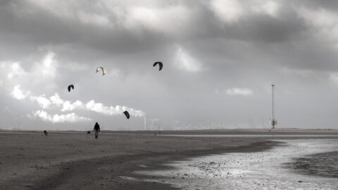Een eenzaam strand met vijf vliegers in de lucht, bewolkte hemel en industrieel landschap op de achtergrond.