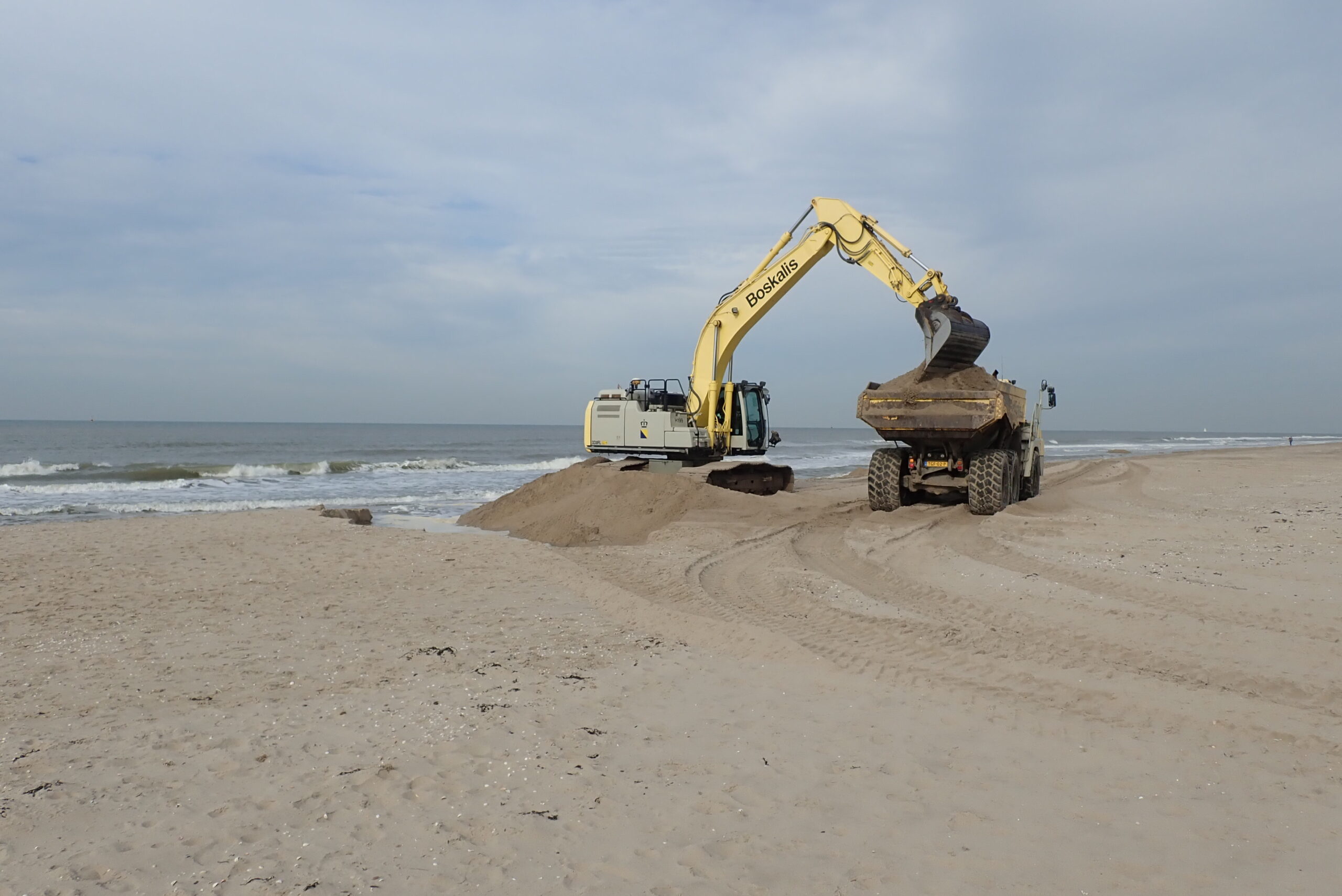 Graafmachine laadt zand in een vrachtwagen aan een rustige kustlijn.