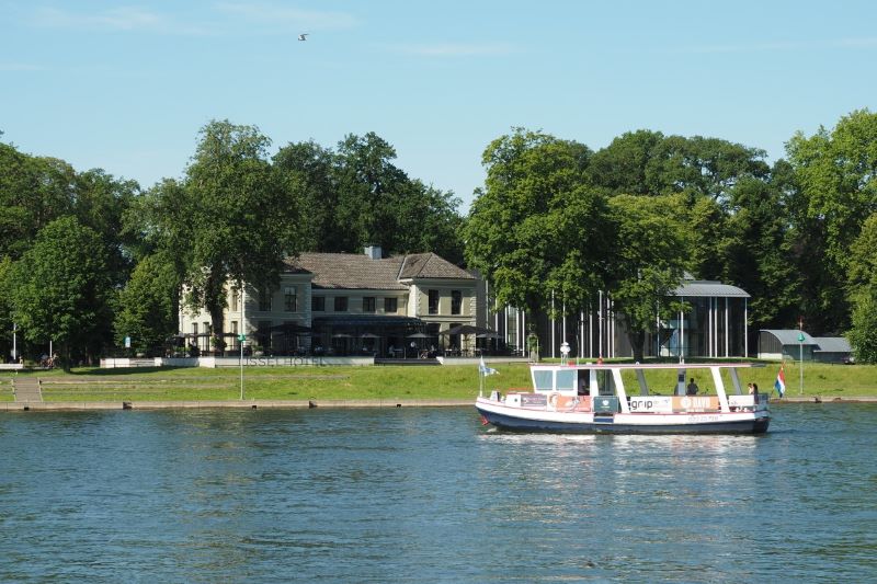 Een boot vaart op een rivier voor een gebouw te midden van groene bomen.