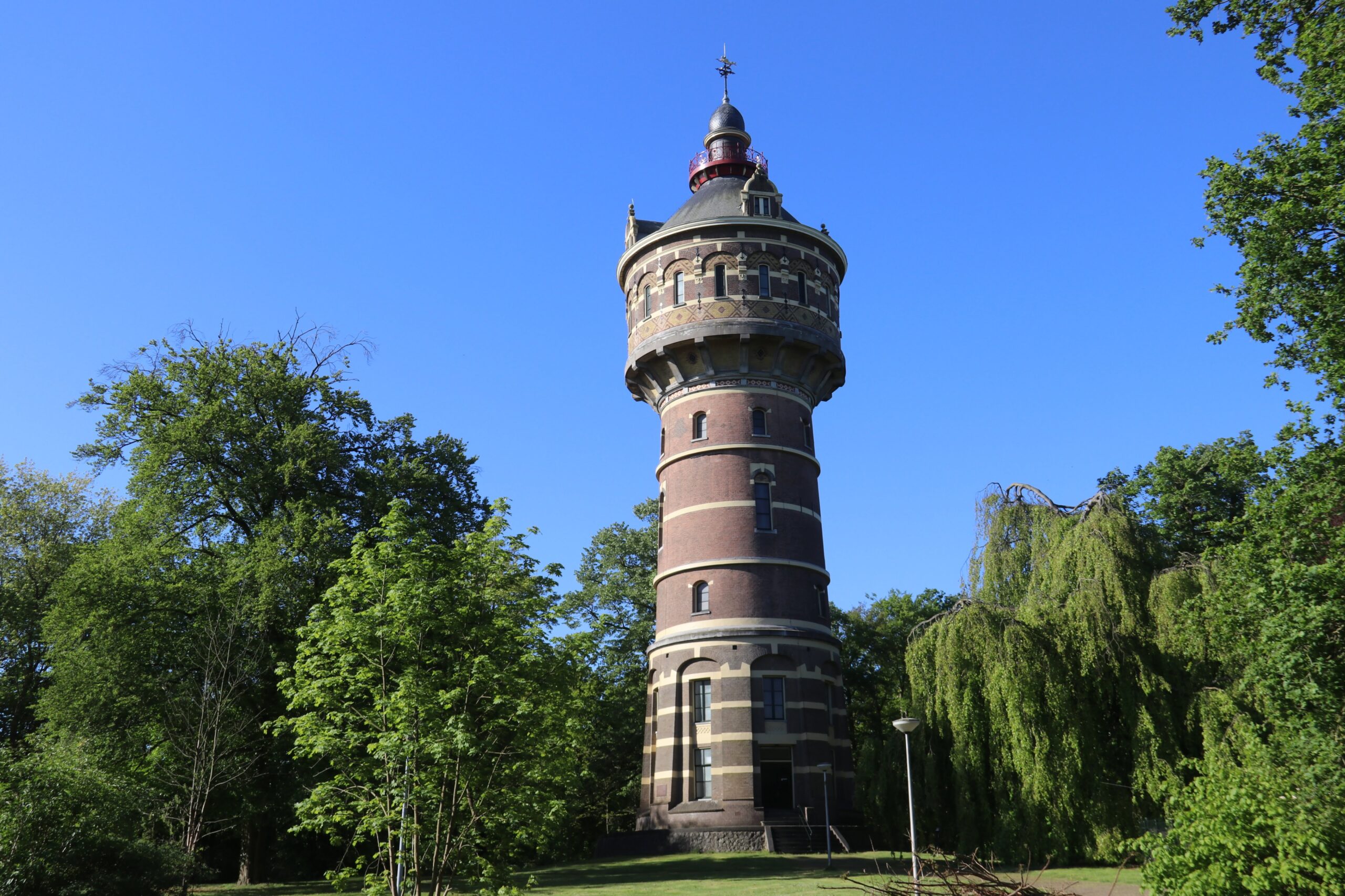 Een oude stenen watertoren omgeven door bomen onder een heldere blauwe lucht.