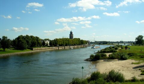 Rivier met groene oevers, bomen, een brug en toren in de verte onder een blauwe lucht met wolken.