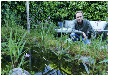 Man in een tuin met een vijver en planten, zit gehurkt naast een bank in een groene omgeving.