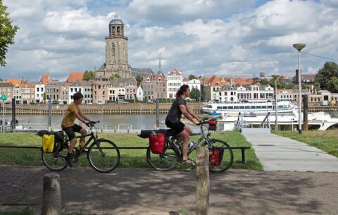 Twee fietsers langs een rivier met historische gebouwen en een kerktoren op de achtergrond.